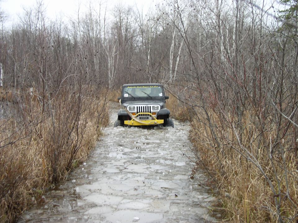 Black Jeep Wrangler with yellow bumper driving on a muddy, icy trail through bare trees
