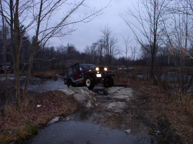 Jeep Wrangler driving through shallow water and over rocks on a trail. Another vehicle is visible in the background