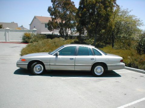 1994 Ford Crown Victoria - A silver sedan is parked on a paved surface with trees and houses in the background.