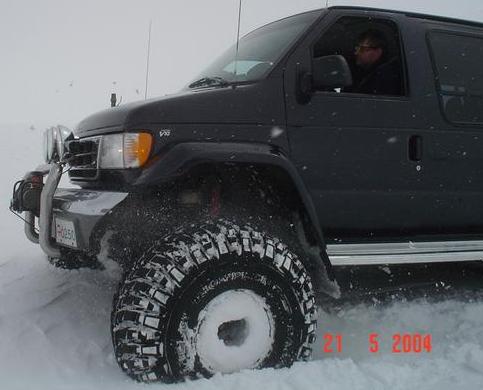 Lifted black Ford van on large off-road tires driving through deep snow, front 3/4 view.