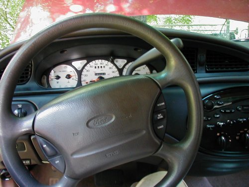 Dashboard of a 1997 Ford Taurus with a gray steering wheel and white gauges. Red hood visible above
