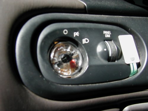 Close-up of a car's interior dashboard showing a headlight switch with symbols for off, parking lights, and headlights, next to a panel dimmer dial