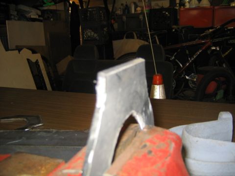 1978 Ford Bronco - Close-up of a bare metal fabricated bracket or arm clamped in the foreground, set on a wood-grain workbench in a cluttered garage. Behind i…