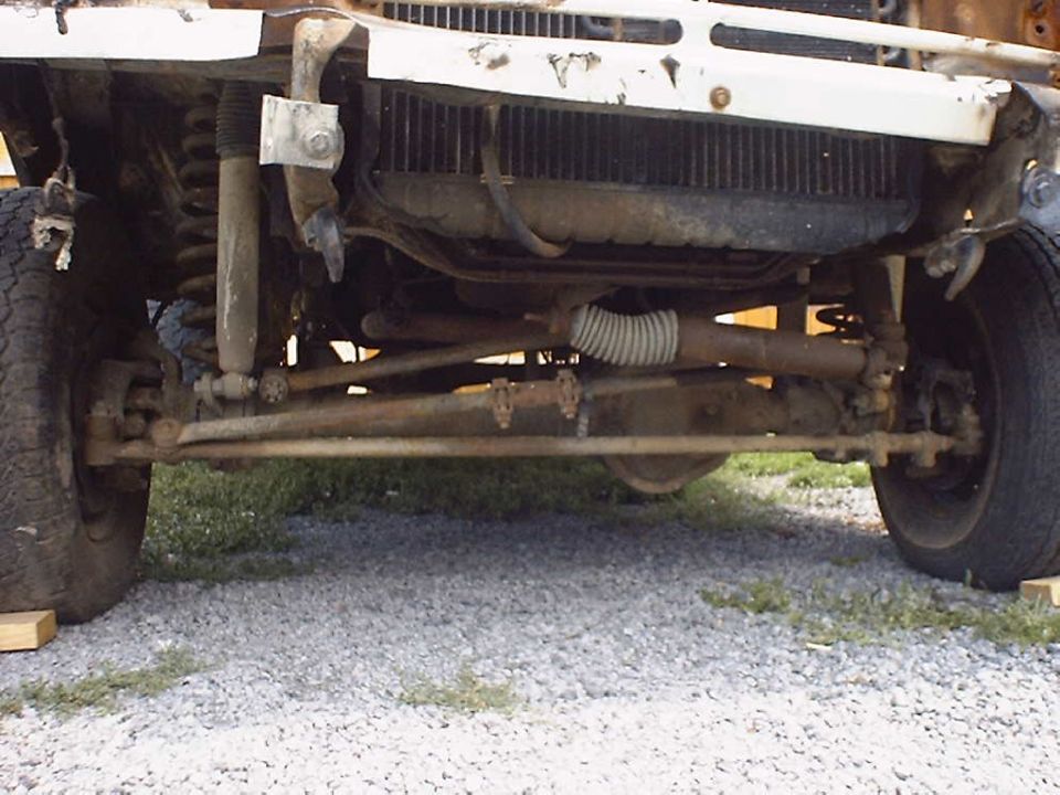 White vehicle's front suspension and grille, showing rust and damage. The vehicle is elevated on wooden blocks