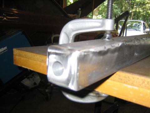 1978 Ford Bronco - Close-up of a fabricated steel tube component with a threaded/bored hole on its end face, held in a C-clamp on a plywood-topped workbench.…