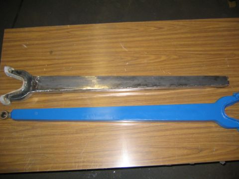 1978 Ford Bronco - Two fabricated suspension radius arms lying on a wood-grain surface, one bare welded steel and one painted blue, each with a forked yoke en…