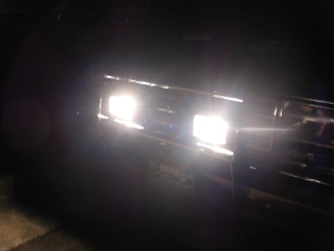 1988 Ford Bronco - The headlights of a dark SUV illuminate the night, with a license plate partially visible on the front grille.