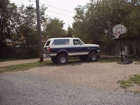 1988 Ford Bronco - Blue-and-white two-tone Ford Bronco with a white removable hardtop parked side-on in a gravel driveway, lifted on oversized mud tires and c…