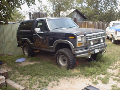 1988 Ford Bronco - A black Ford Bronco with significant rust damage sits in a grassy yard next to another vehicle.