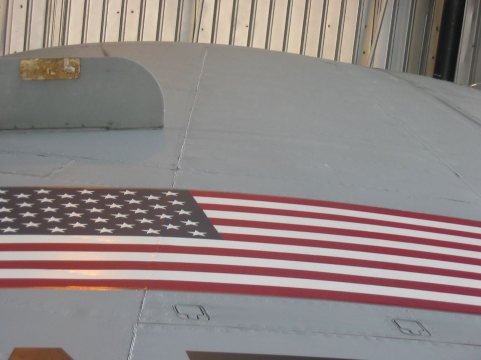 Close-up of an aircraft's gray fuselage with a large American flag decal and a Supermotors.net watermark