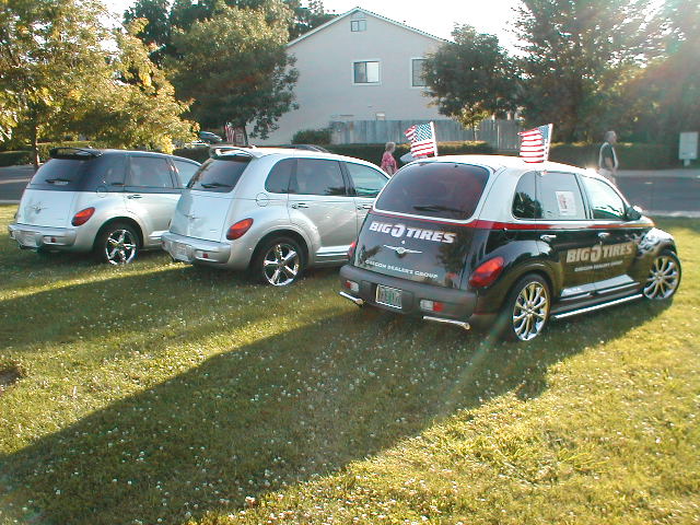 Three PT Cruisers on grass, two silver and one black with Big O Tires graphics and U.S. flags.