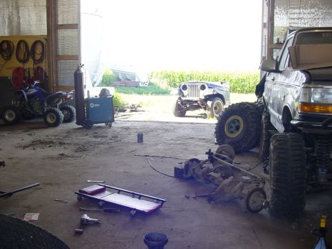 1992 Ford Bronco - Garage scene with a lifted Ford truck, a Jeep, an ATV, and parts, suggesting ongoing vehicle work.