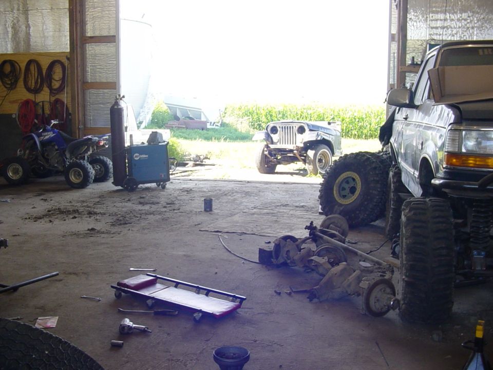 Garage interior with a lifted Ford truck, a disassembled Jeep, an ATV, and various automotive parts scattered around