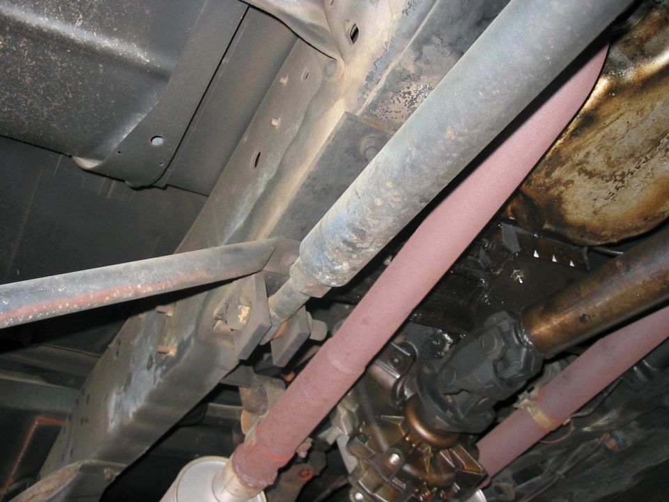 Underside view of a vehicle's frame, driveshaft, and exhaust system. Some rust is visible on the exhaust pipes