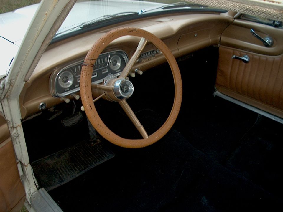 Tan interior of an old car with large thin steering wheel, dash gauges and bare floor