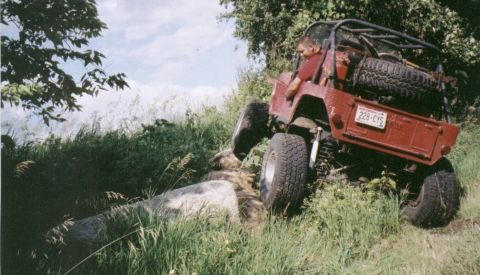 1980 Ford Bronco - A red off-road vehicle with a roll cage and spare tire tackles a challenging rocky path.