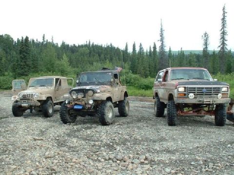 1985 Ford Bronco - Three off-road vehicles, including a Ford Bronco, are parked on a gravelly surface with a forest in the background.