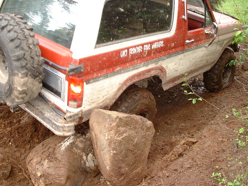Muddy red-and-white Ford Bronco crawling over boulders, rear 3/4 view