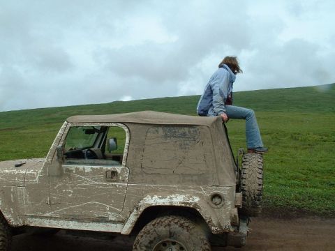 1985 Ford Bronco - A person sits atop a mud-splattered Jeep Wrangler in a grassy, hilly landscape under a cloudy sky.