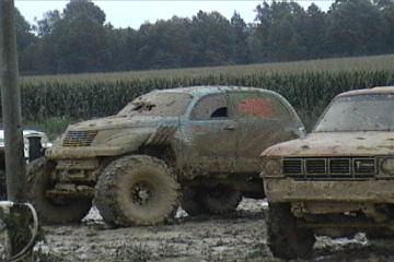 1989 Ford Bronco - Two off-road vehicles are covered in mud, parked next to a cornfield.