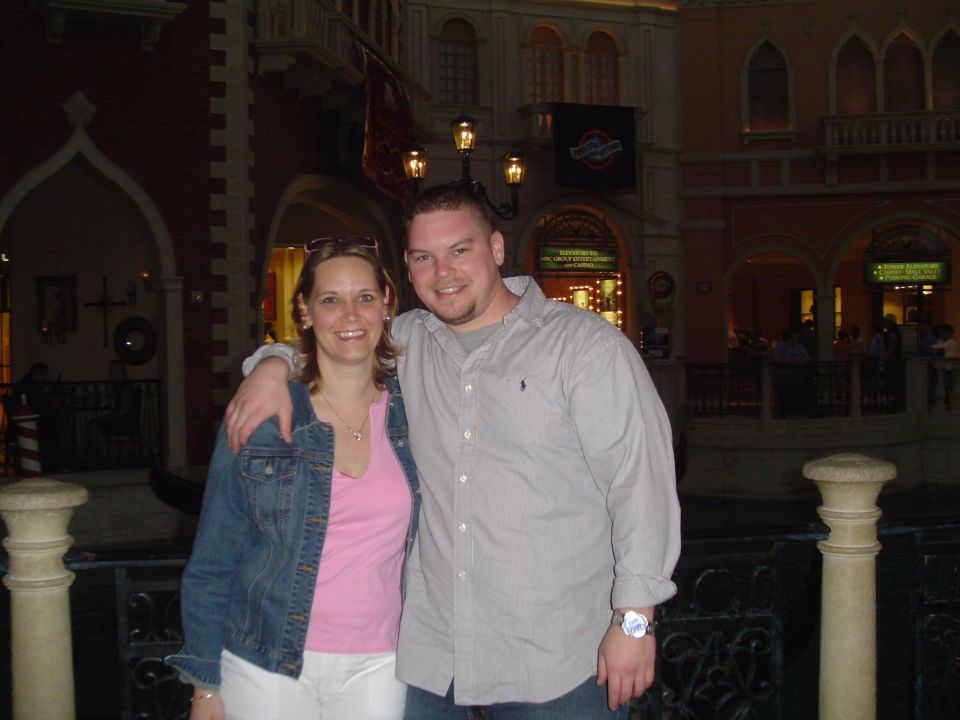 Two people posing together in front of an indoor Venetian-style canal and arched facades