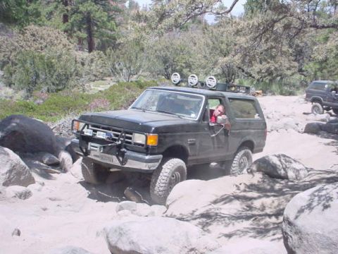 1988 Ford Bronco - A black Ford Bronco is shown navigating a challenging off-road trail with large rocks and sand.
