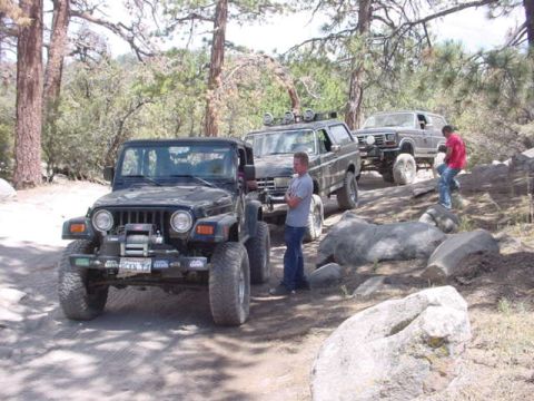 1988 Ford Bronco - Three off-road vehicles navigate a challenging path through a wooded, rocky area.