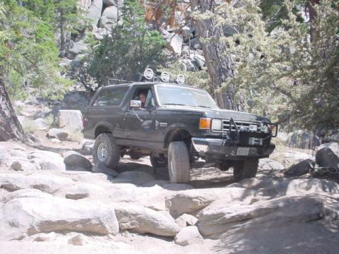 1988 Ford Bronco - A Ford Bronco navigates a rocky off-road trail surrounded by trees and boulders.