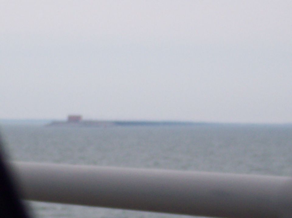 Water with a distant red structure on a hazy horizon, foreground shows a blurred gray bar