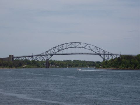 1990 Ford Bronco - A large steel arch bridge spans a wide waterway under a cloudy sky, with a speed boat and sailboats visible.