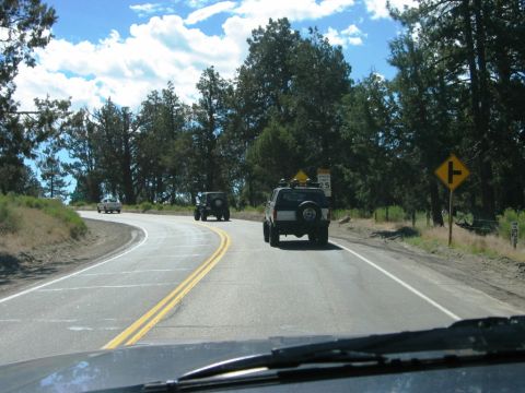 1985 Ford Bronco - Two SUVs travel down a paved road surrounded by trees under a partly cloudy sky.