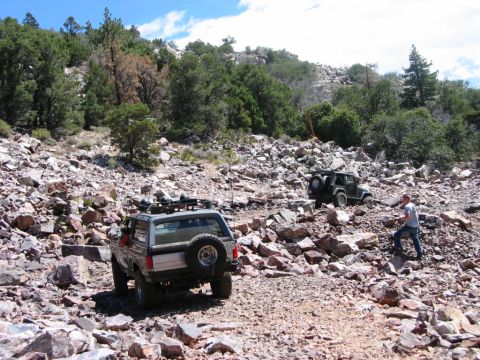 1985 Ford Bronco - Two off-road vehicles navigate a challenging, rocky mountain trail under a partly cloudy sky.