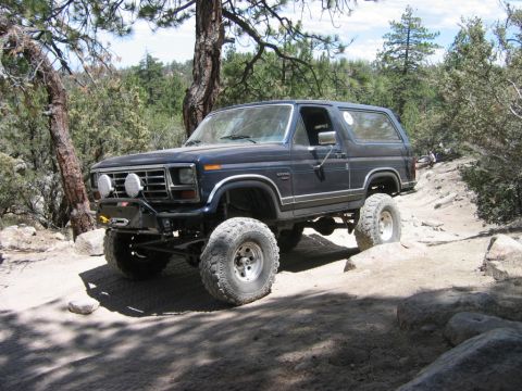 1985 Ford Bronco - A lifted Ford Bronco navigates a rocky, wooded trail.