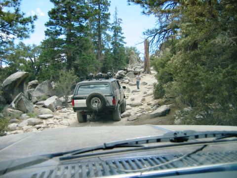 1985 Ford Bronco - An SUV navigates a challenging rocky path through a forest, with another vehicle's dashboard visible in the foreground.