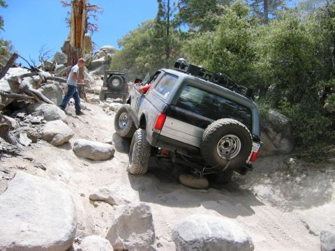 1985 Ford Bronco - Two SUVs tackle a challenging off-road course on a sunny day.