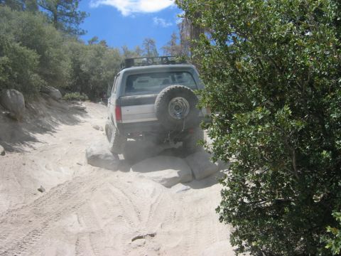 1985 Ford Bronco - A gray Ford Bronco navigates a challenging off-road path with rocks and sand.