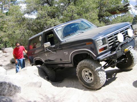 1985 Ford Bronco - A dark 1985 Ford Bronco navigates a challenging off-road course with a person standing nearby.