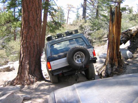 1985 Ford Bronco - An SUV is shown off-roading on a rocky trail, with a spare tire on the back and a roof rack.