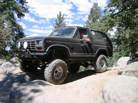 1985 Ford Bronco - A modified black Ford Bronco tackles a challenging off-road course on a sunny day.