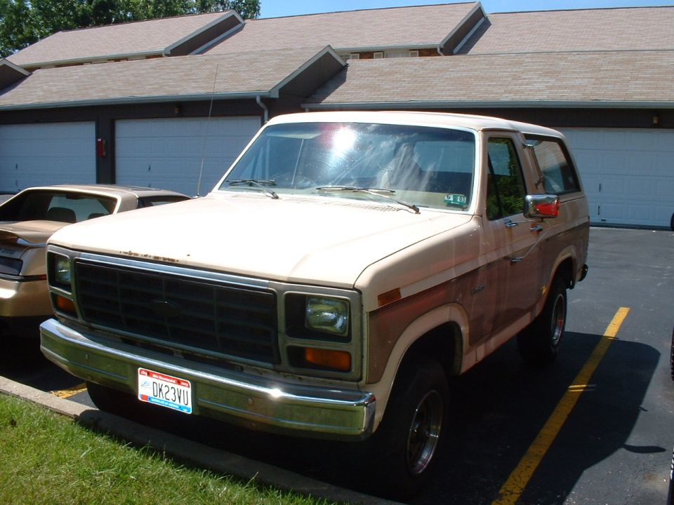Tan and brown Ford Bronco parked in front of garages, with another car to its left
