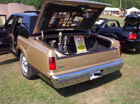 1991 Ford Crown Victoria LTD - Gold sedan's trunk is open, displaying trophies and a reserved sign, with another car behind.