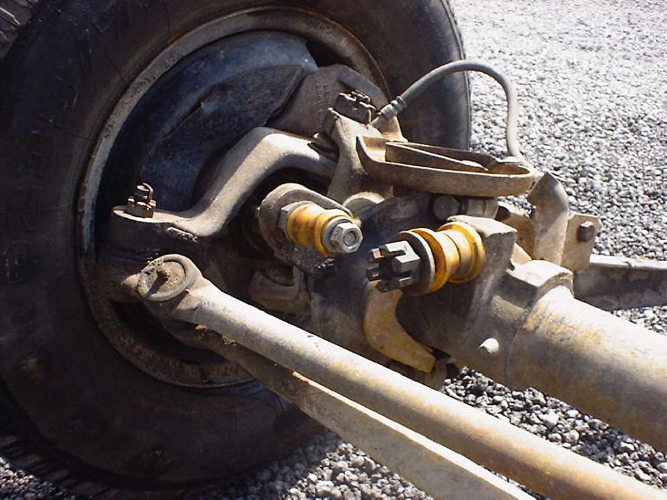 Close-up of a vehicle's front axle and suspension with a tire visible. Rusty metal parts and yellow bushings are prominent