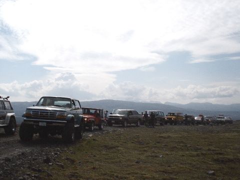 1996 Ford Bronco - A line of off-road vehicles, including a Ford F-Series truck, parked on a grassy, rocky terrain under a cloudy sky.