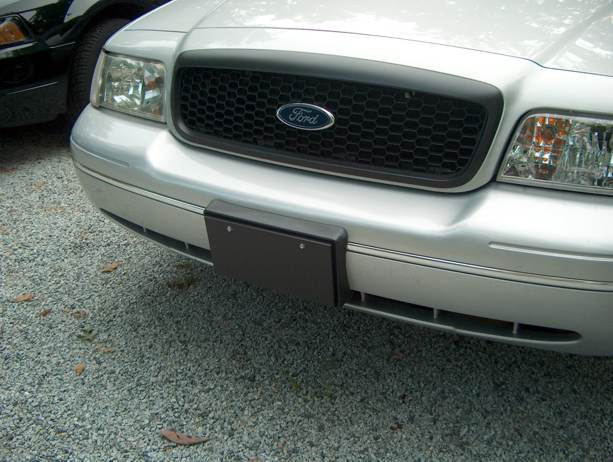 Front grille and bumper of a silver Ford Crown Victoria with blank black license plate bracket, parked on gravel