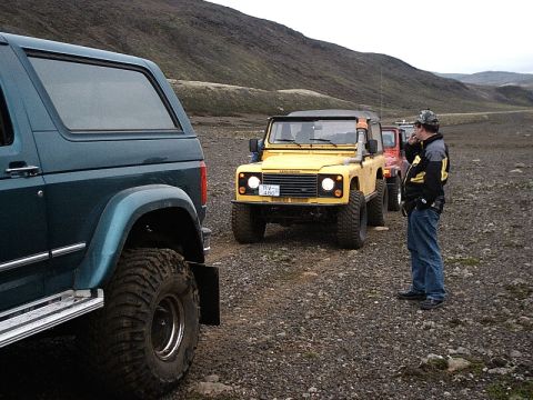 1996 Ford Bronco - A teal SUV is parked in the foreground, with a yellow Land Rover Defender and other vehicles lined up behind it on a gravel road.