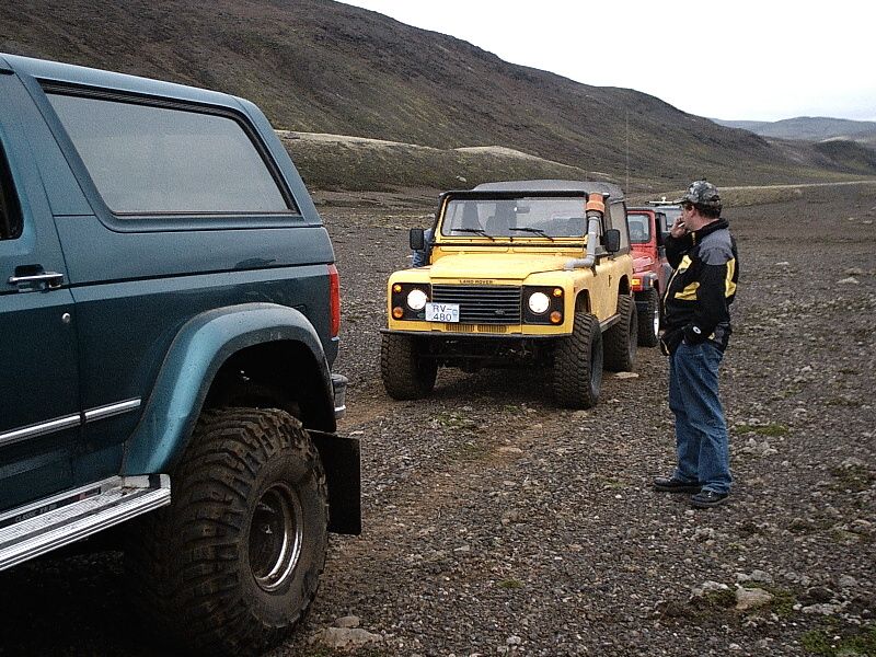 Teal Ford Bronco parked beside a yellow Land Rover on gravel in a barren hilly terrain