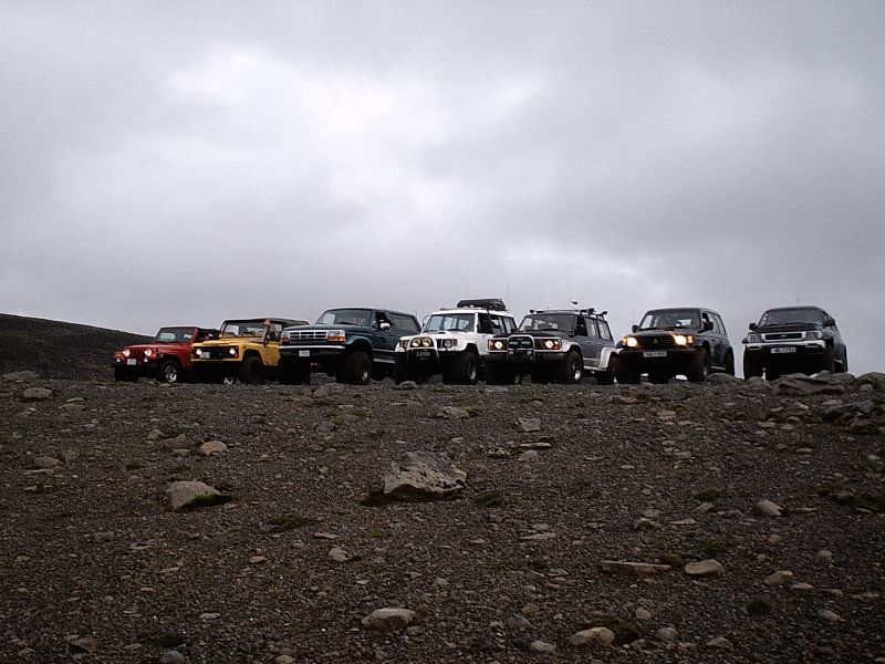 Row of seven lifted 4x4 SUVs parked on rocky terrain under a cloudy sky