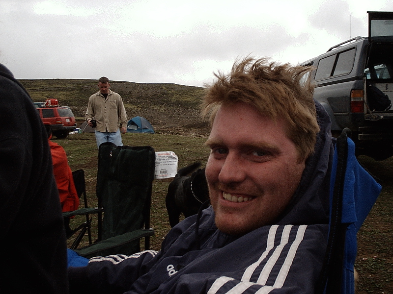 Man in blue jacket at campsite with dark SUV and red vehicle in background