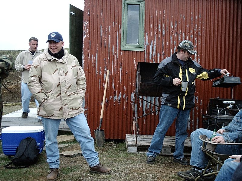 Men standing outdoors by a rusty red metal shed with a grill and cooler