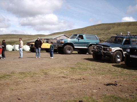 1996 Ford Bronco - Several off-road vehicles are parked in a gravel lot with a building and hills in the background.